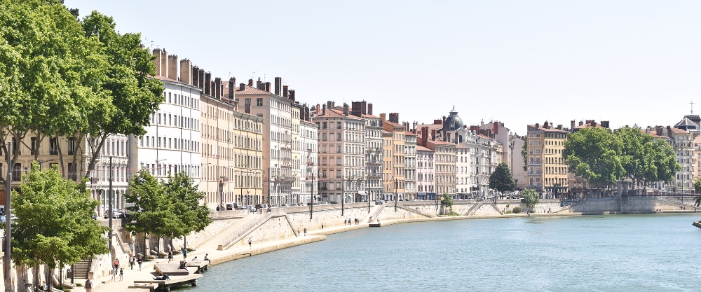 Pedestrian walkway along the river in Lyon with classic residential buildings and trees lining the shore.